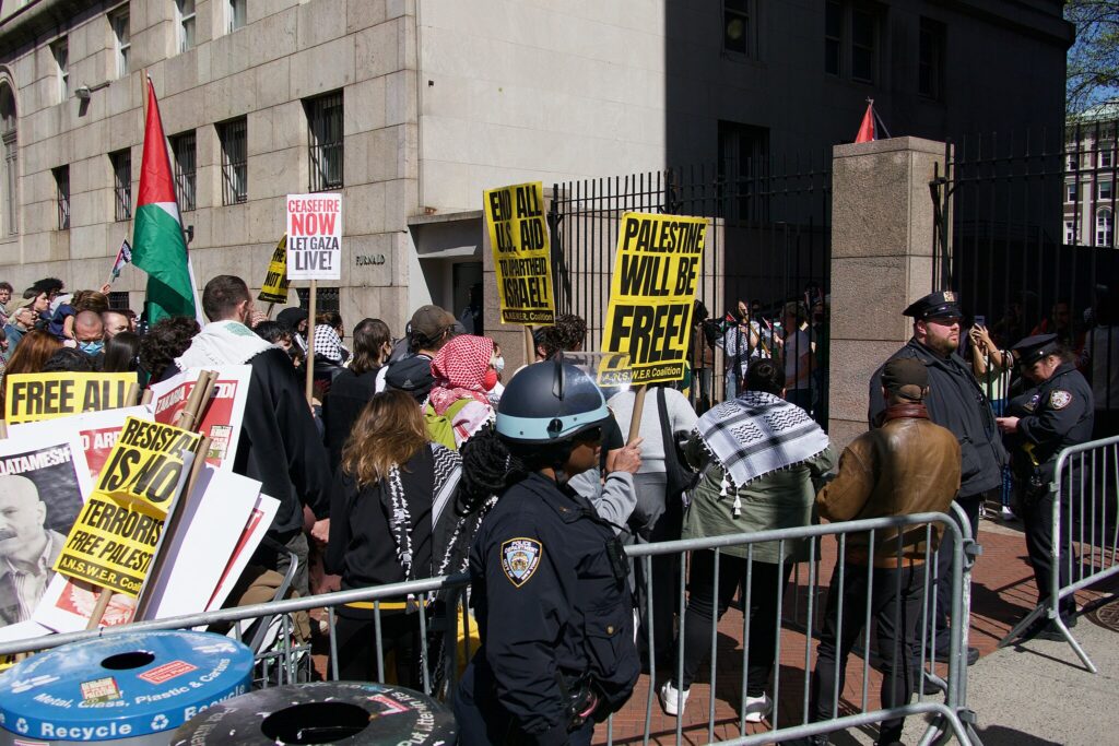 Pro-Palestine protestors stand on a sidewalk outside of a gated entrance to Columbia University. A handful of police officers stand around the crowd, who are hemmed in by moveable steel gates.